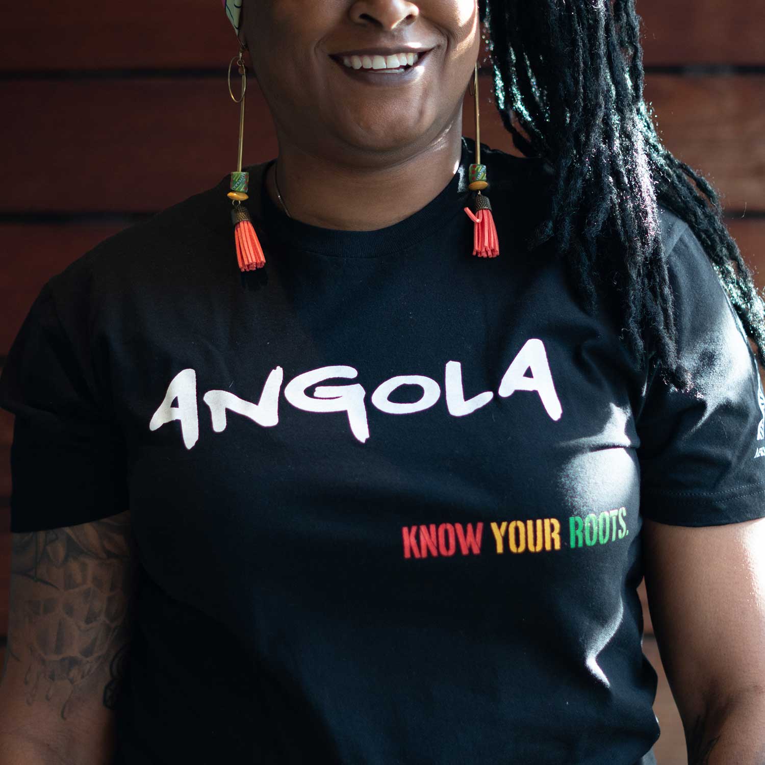 African American woman wearing the Angola T-shirt with "Angola" written in white and "Know Your Roots" written in African colors 