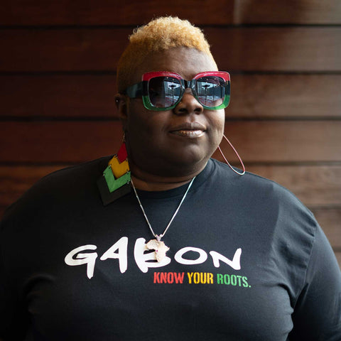 African American Woman wearing a Black T-shirt that says "Gabon" in white and "Know Your Roots" in African colors.