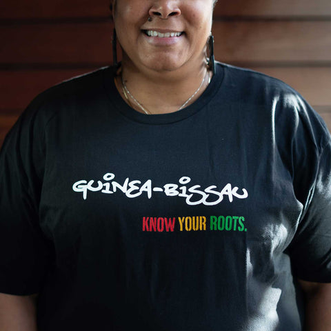 African American Woman wearing a black T-shirt that says "Guinea-Bissau" in white and "Know Your Roots" in African colors. 