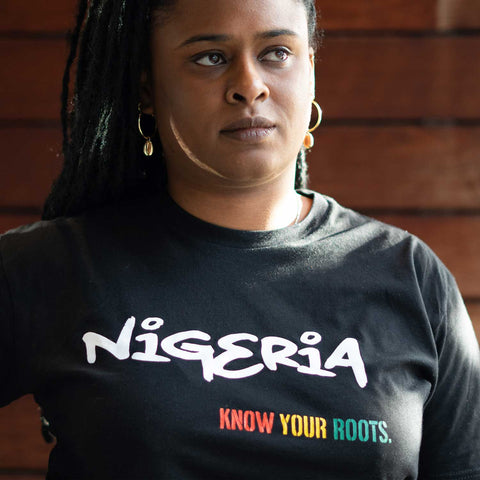 African American woman wearing a black t-shirt that says "Nigeria" in white and "Know Your Roots" in African colors. 
